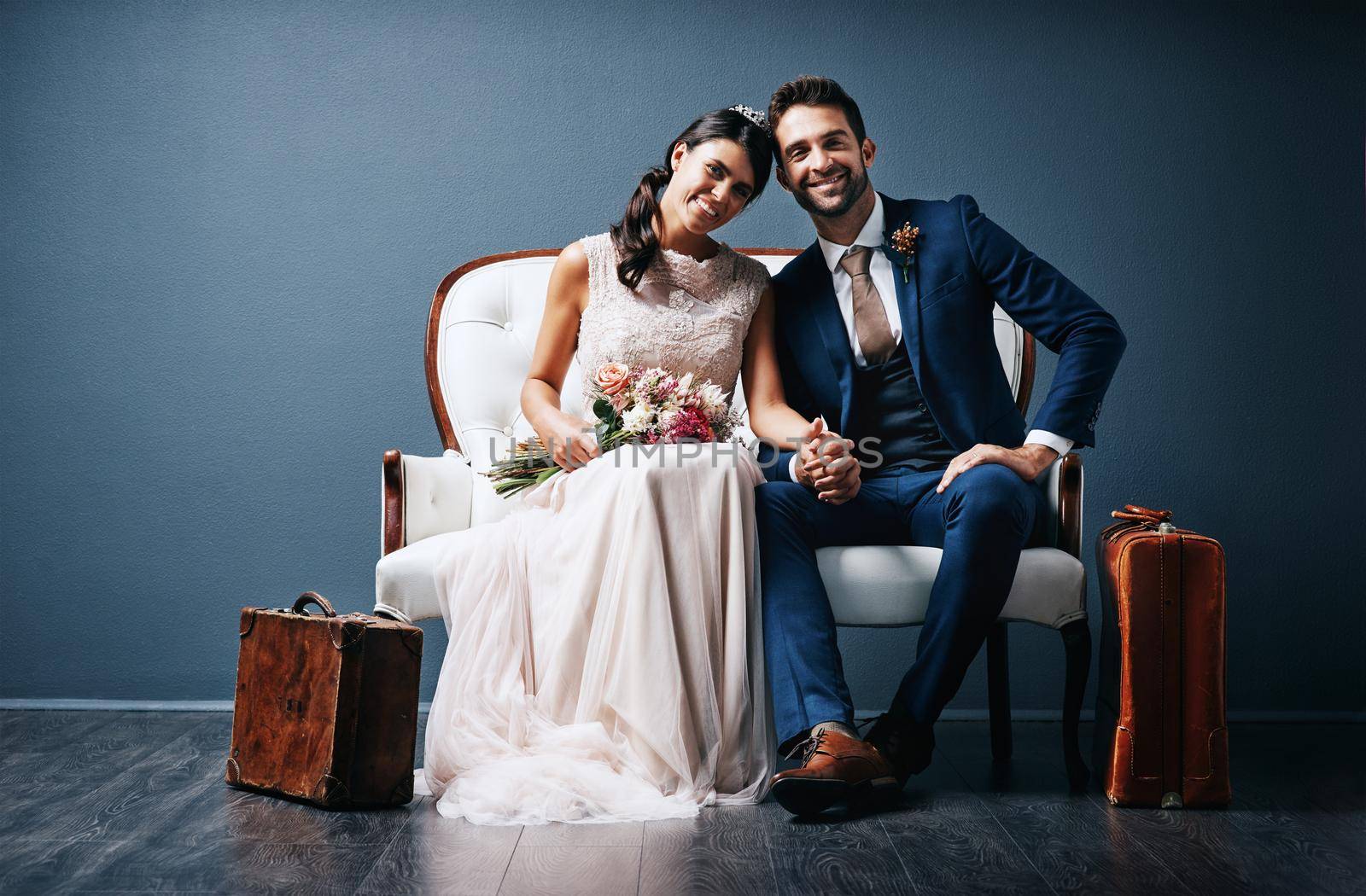 Packed up and ready for our adventure. Studio shot of a newly married young couple sitting together on a couch against a gray background. by YuriArcurs