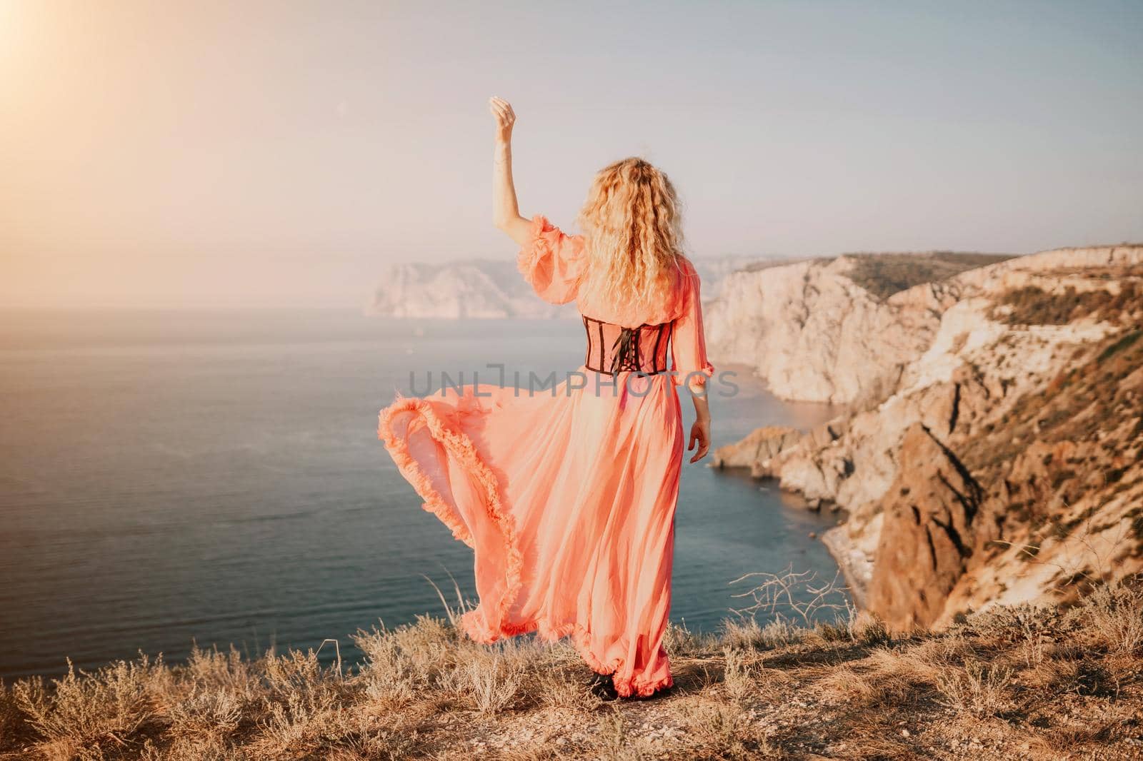 Side view a Young beautiful sensual woman in a red long dress posing on a volcanic rock high above the sea during sunset. Girl on the nature on blue sky background. Fashion photo