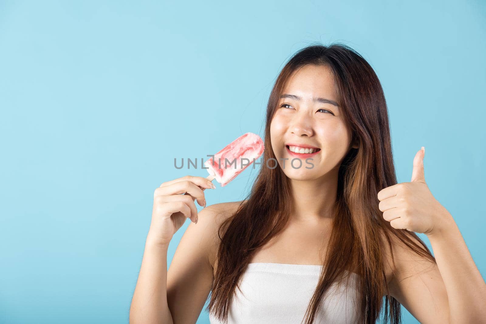 Sweet tasty frozen dessert icecream on summer time. Happy Asian young beautiful woman holding delicious ice cream wood stick mixed fruit flavor and show thumb up good sign, isolated on blue background