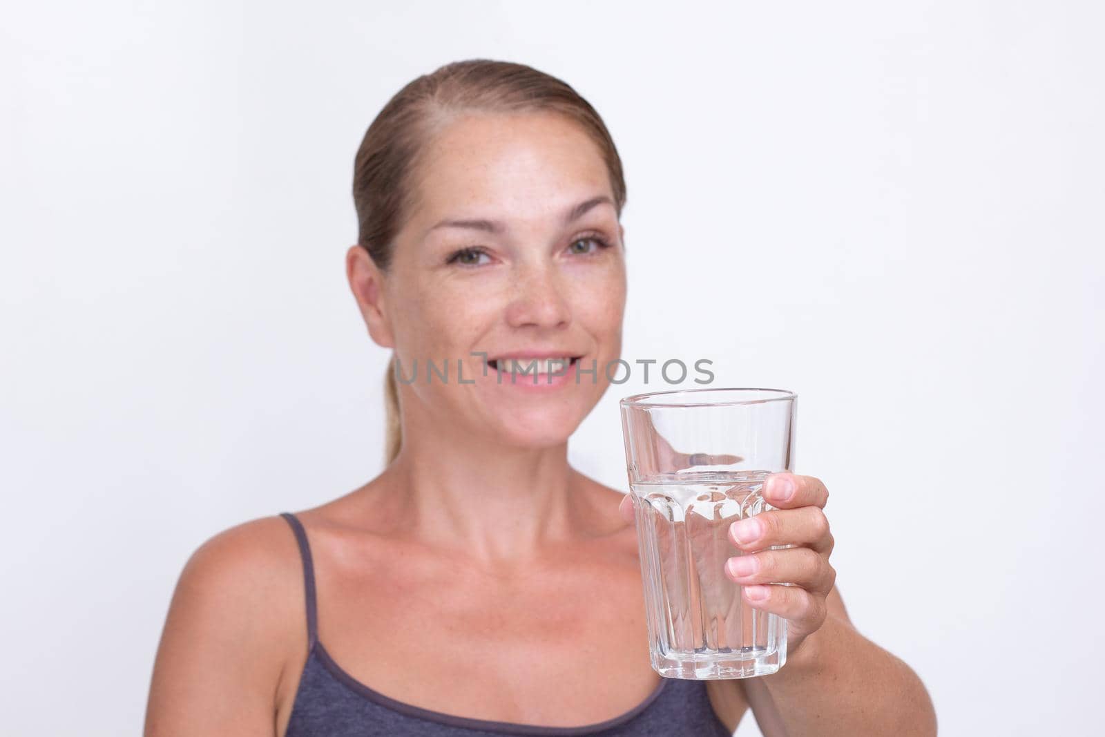 Caucasian beautiful smiling middle aged woman holding glass of pure water on white background looking at camera. Focus on glass