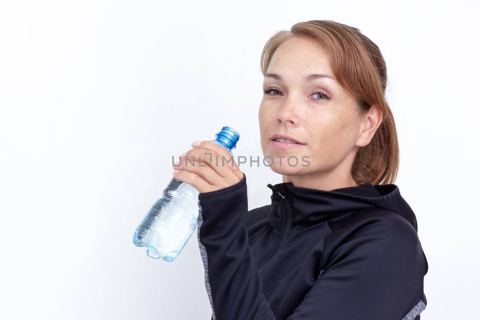 Portrait of caucasian smiling middle aged woman holding plastic bottle of water near mouth to drink on white background looking at camera