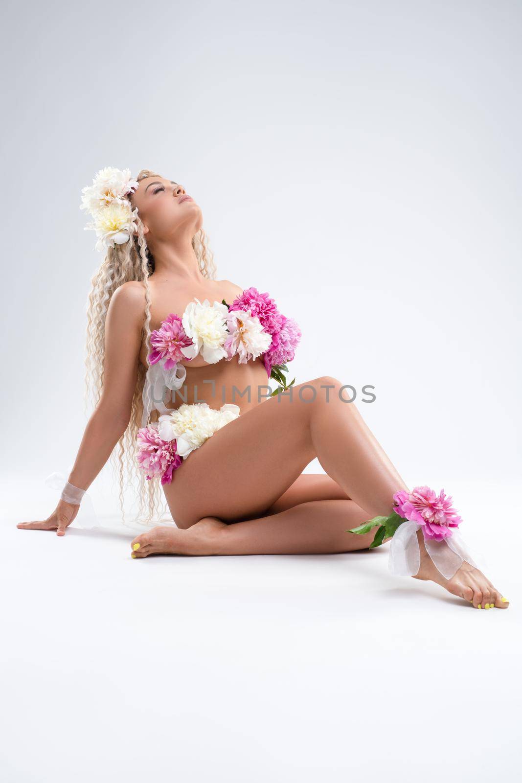 Full body of sensual female model with long blond curly hair and bunch of fresh flowers on naked body sitting on floor against white background in studio
