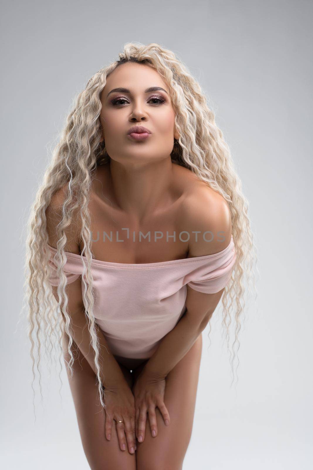 Self assured alluring young female model with long curly blond hair and bare shoulders in bodysuit leaning forward and pouting lips against gray background