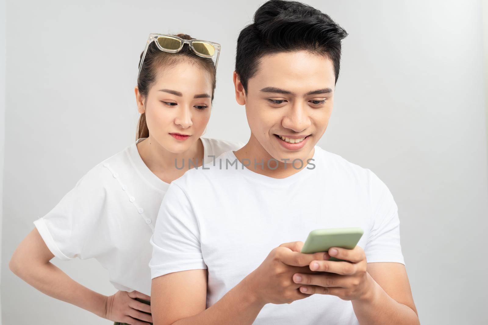 Curious boyfriend is spying his lovers smartphone. They are wearing casual shirts, standing isolated on white background