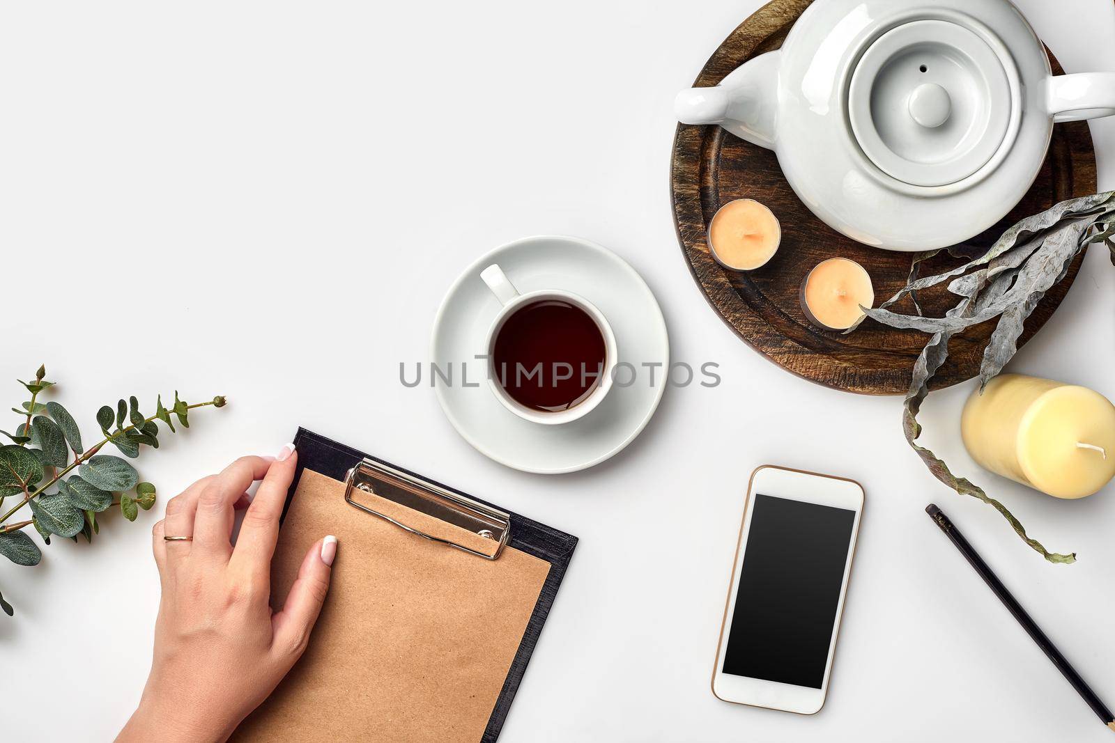 Young woman right hand writing on blank notebook on white table with tea cup, smartphone, and kettle beside in morning time. Top view. Copy space.