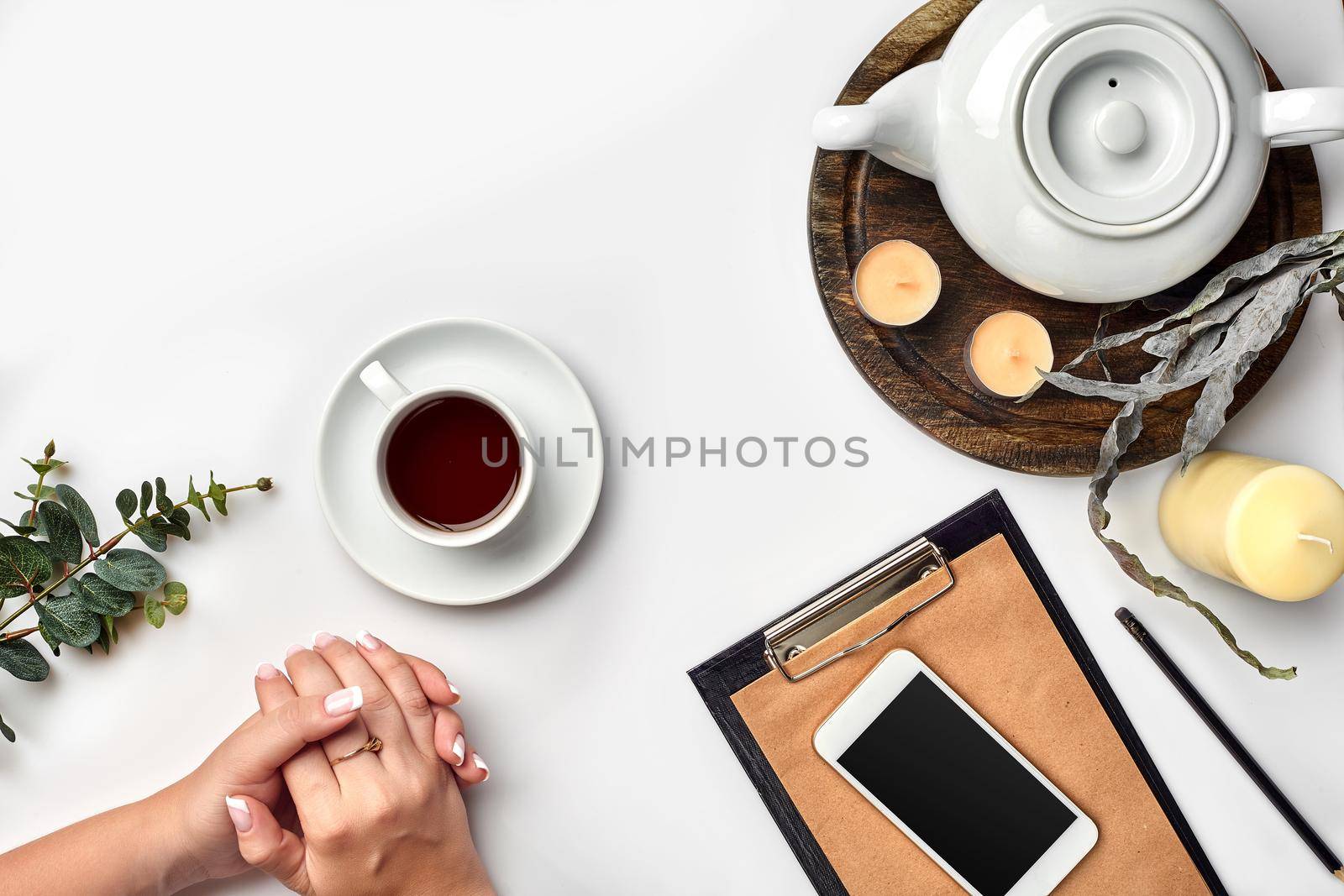 Young woman right hand writing on blank notebook on white table with tea cup, smartphone, and kettle beside in morning time. Top view. Copy space.