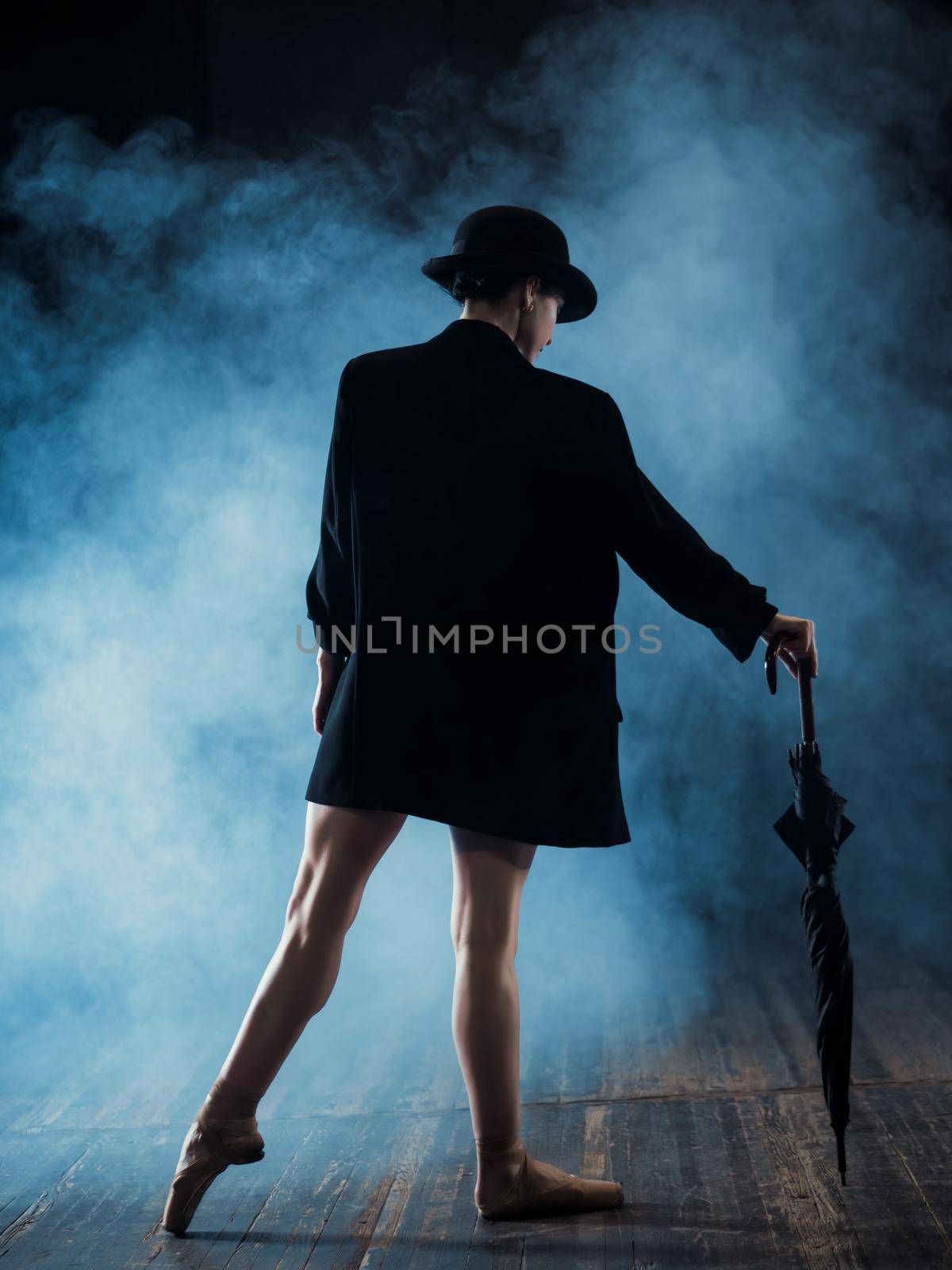 Ballerina in black long jacket and sexy bodysuit dancing on stage with magic light and smoke. Young attractive dancer in ballet shoes pointe with umbrella.