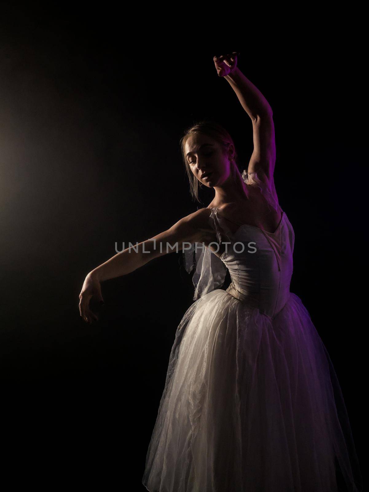 Ballerina is practicing her moves in dark studio. Young girl dancing with air white dress tutu, spinning around and smiling. Gracefulness and tenderness in every movement