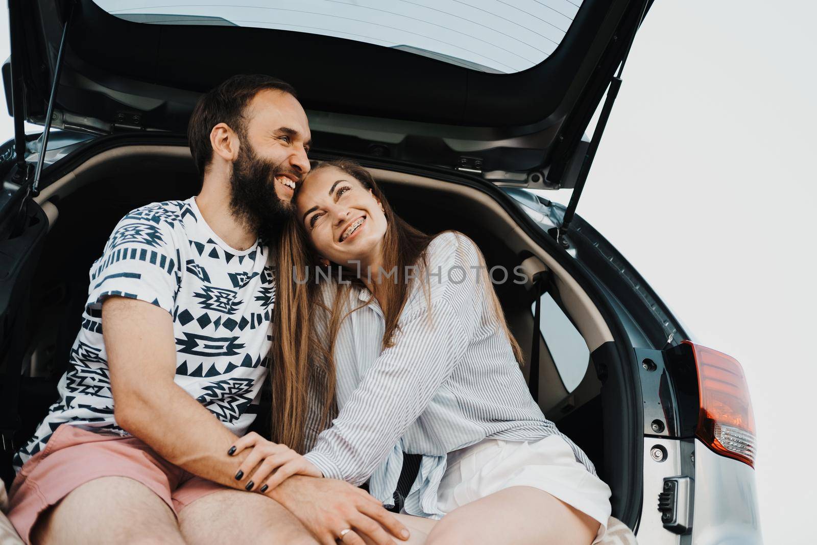 Happy middle-aged couple making stop while on a road trip, caucasian cheerful woman and man sitting in trunk of SUV car and smiling