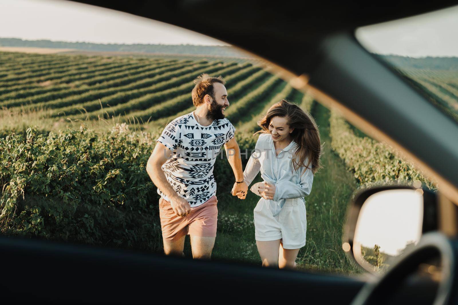 View through car window, happy middle-aged couple running in the beautiful fields, caucasian man and woman enjoying vacation outdoors by Romvy