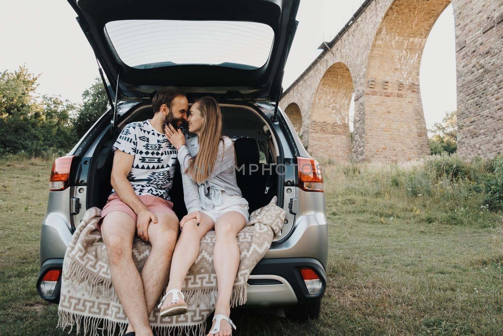 Middle-aged couple sitting inside trunk of SUV car with viaduct bridge on the background, caucasian man and woman enjoying road trip together by Romvy