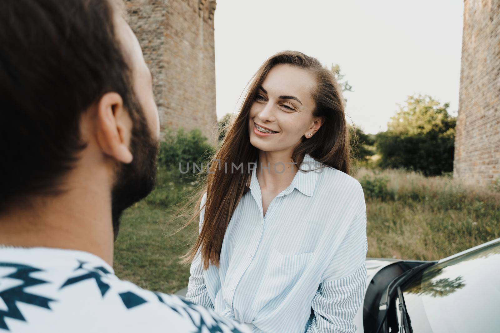 Caucasian woman sitting on hood of car and smiling to her boyfriend, middle-aged couple enjoying road trip together