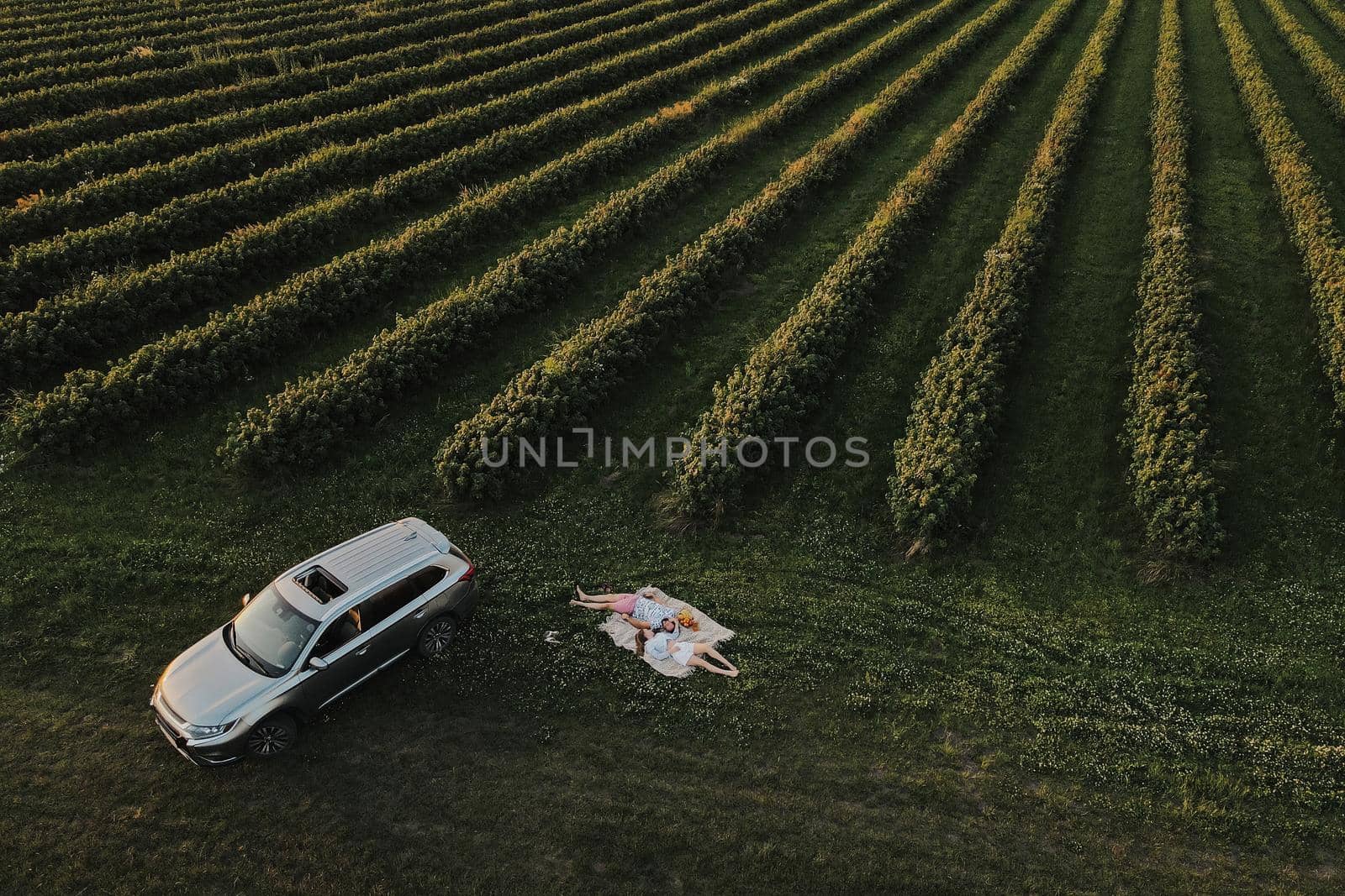 Aerial shot couple enjoying picnic on road trip in the beautiful fields, man and woman lying on blanket near the SUV car at sunset