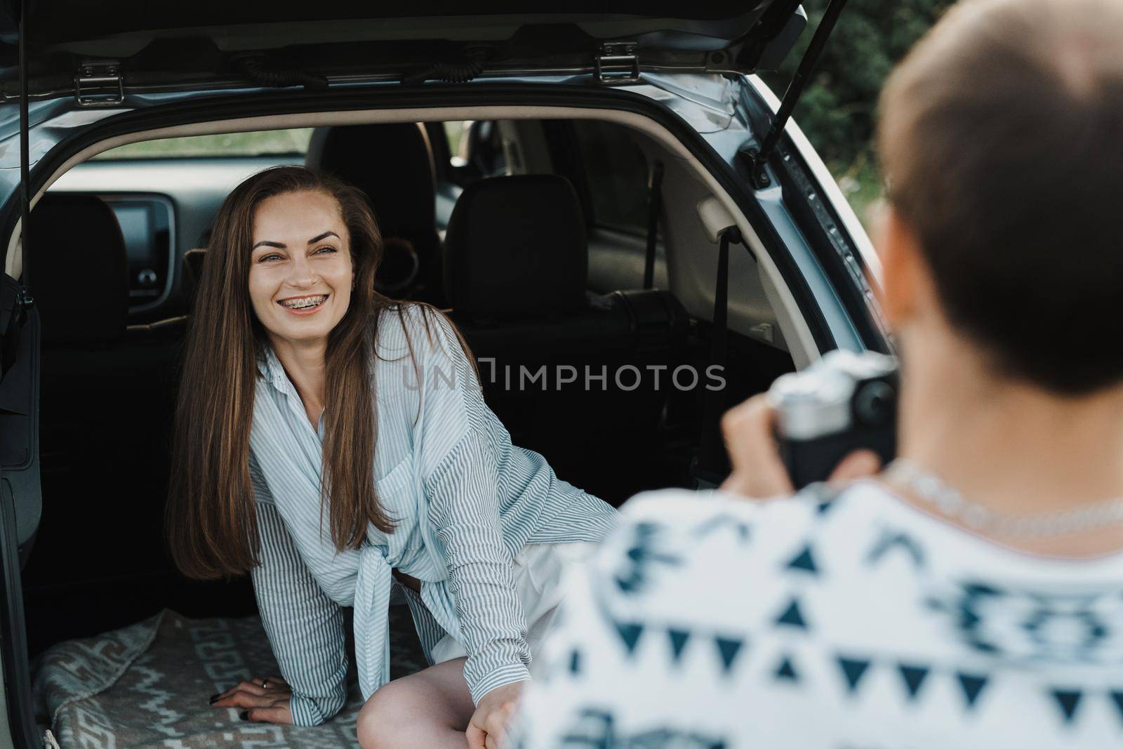 Cheerful caucasian woman sitting in trunk of SUV car while boyfriend photography her on a camera by Romvy