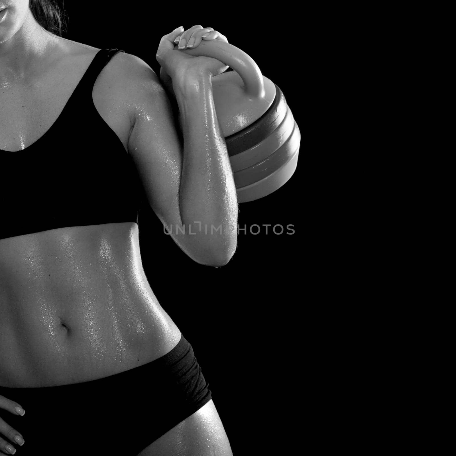 Young sporty girl with muscular body in black lingerie with the sweat on the skin does exercise standing with weights on a black background. Black and white photography.