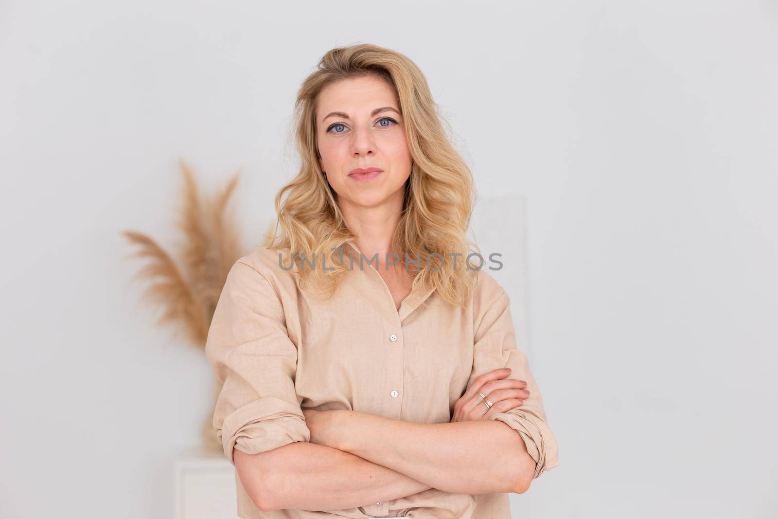 Portrait of a beautiful woman, 30-40 years old, with blond hair, stands in a beige linen shirt and white trousers, in a white room interior. Copy space