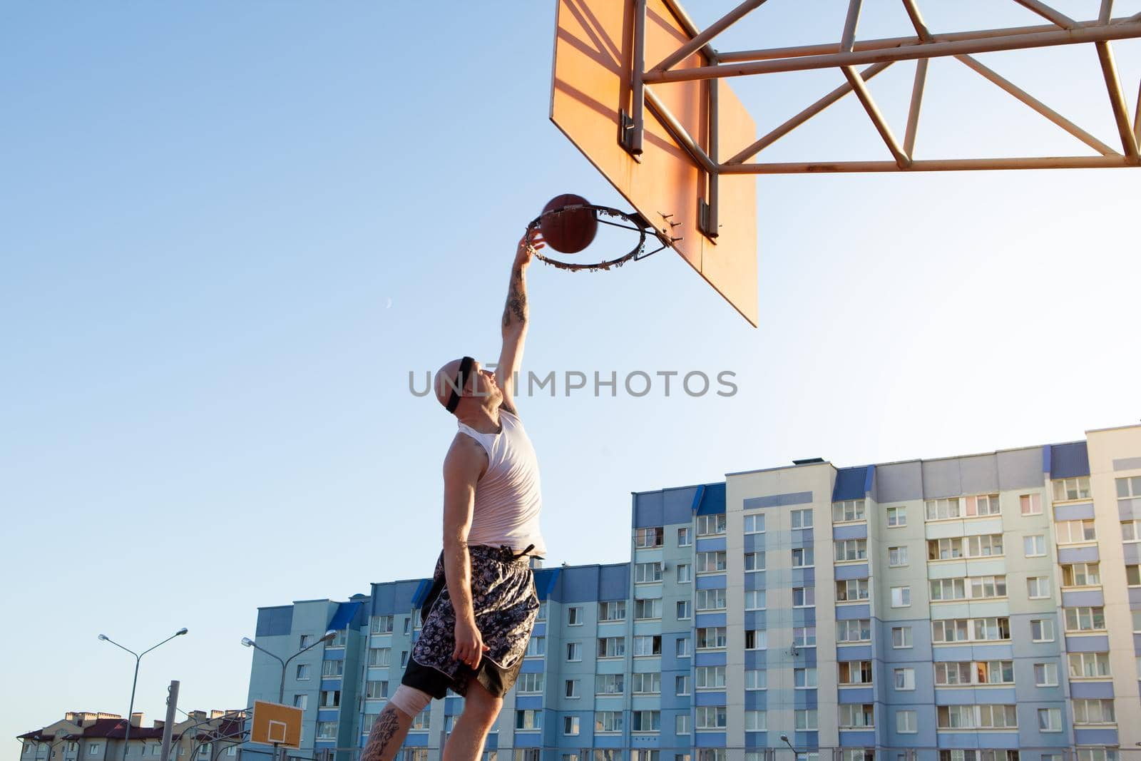 One guy play basketball at district sports ground against a backdrop of high-rise residential buildings. The player throws the ball into the ring in the jump.