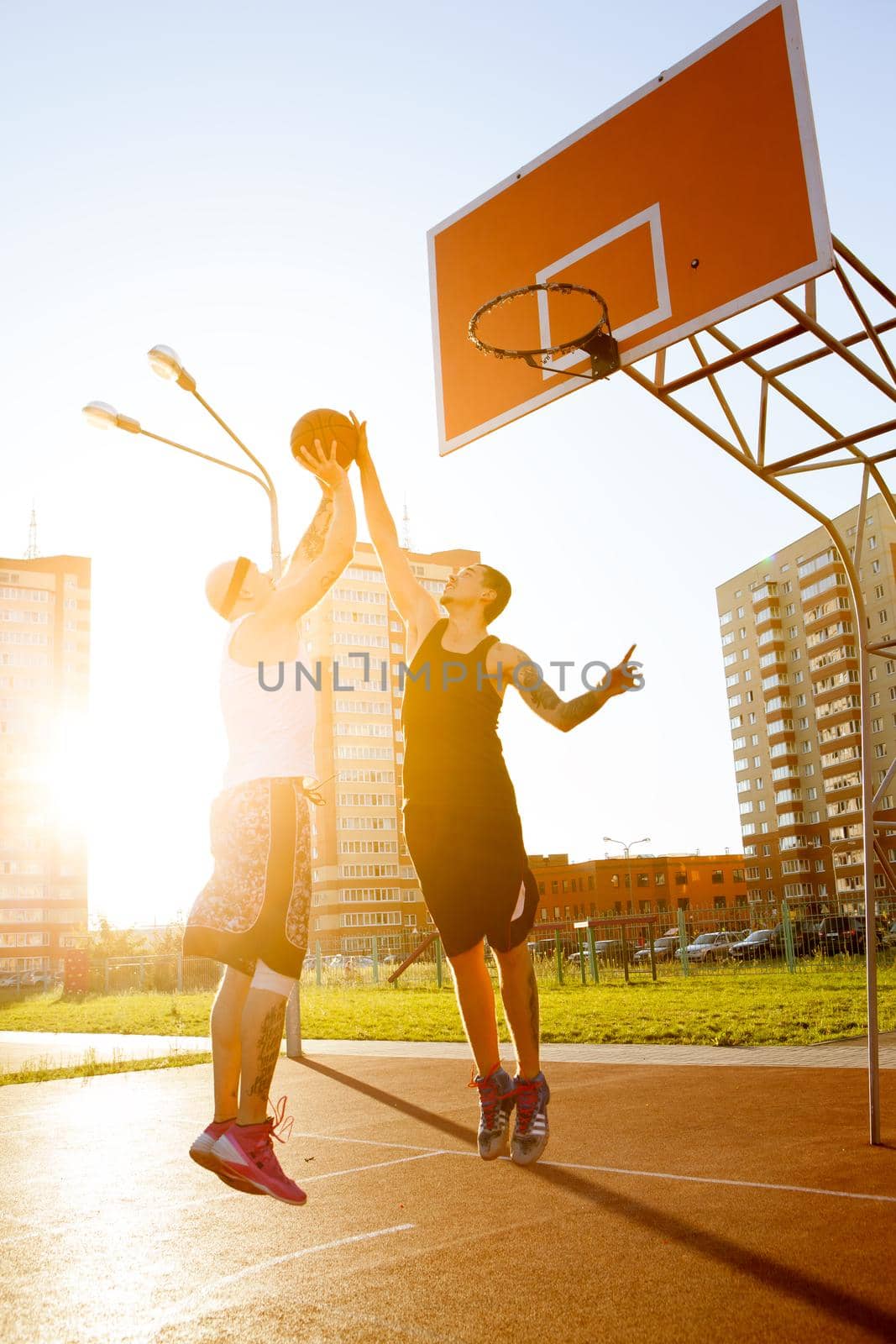 Two stylish guy play basketball at district sports ground against a backdrop of high-rise residential buildings. Solar flare in the frame.