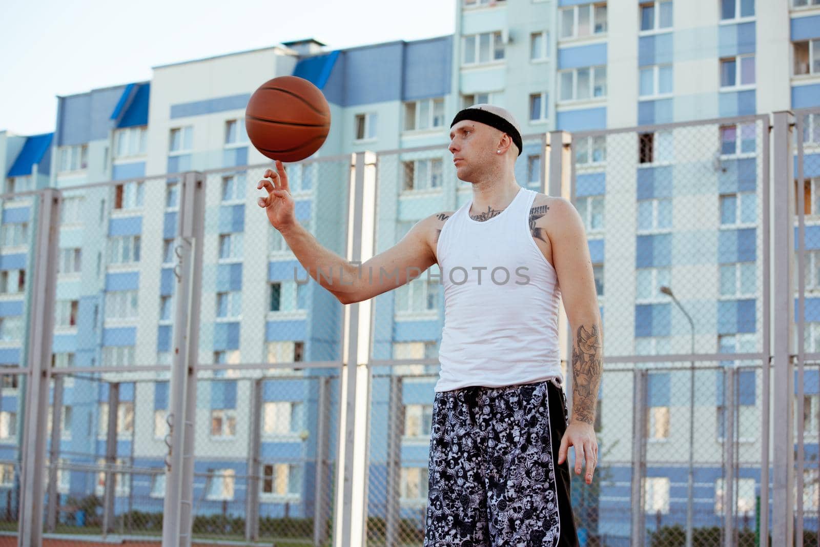 Young man standing on a basketball court the background of houses and spinning the ball his finger. by studiodav