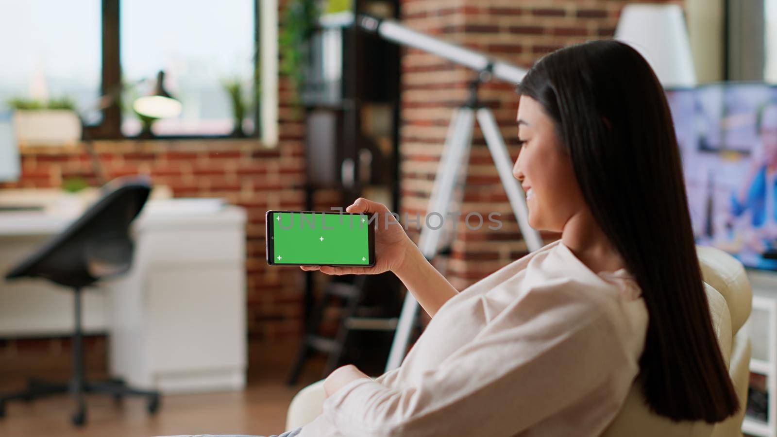 Happy asian person with mobile phone having mockup isolated display while sitting on sofa at home. Smiling woman having smartphone with green screen chroma key background.