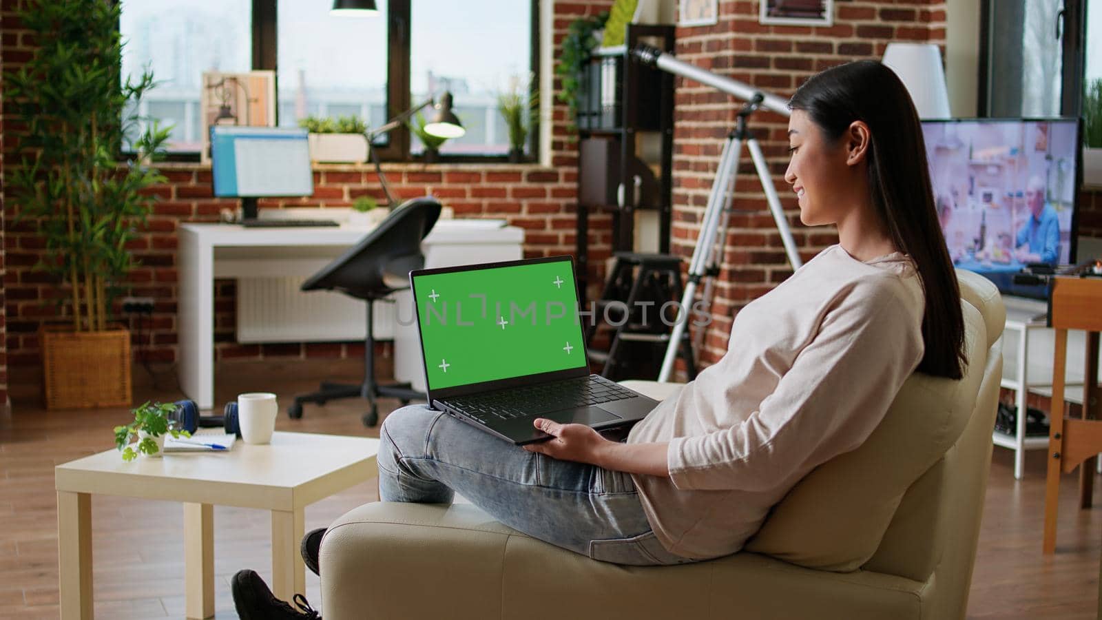 Smiling heartily asian woman using laptop having green screen mockup template display. Happy young adult person sitting on couch at home with portable computer in lap having chroma key background.