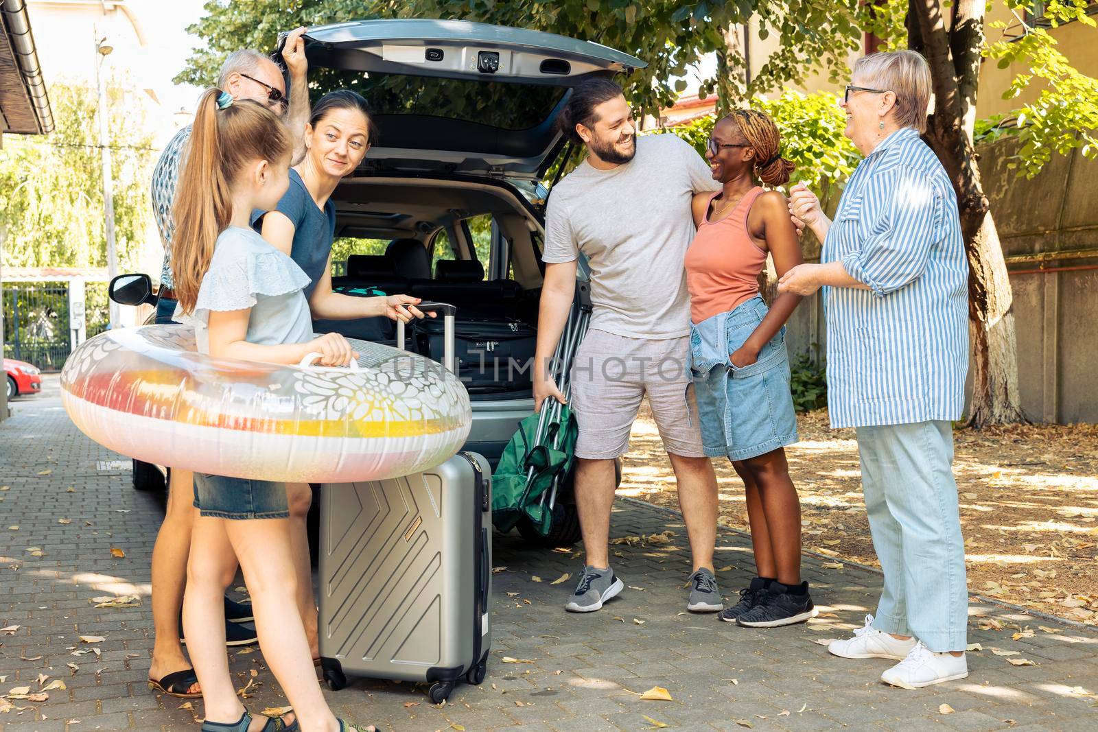 Diverse family and friends travelling on holiday, preparing to leave on summer vacation and loading baggage in car trunk. People sitting in driveway to go to seaside adventure trip.