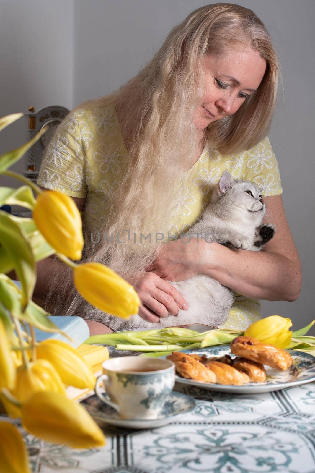 blonde middle-aged hugging a kitten and drinking tea, still life with yellow tulips and cakes a stack of books on the table. High quality photo