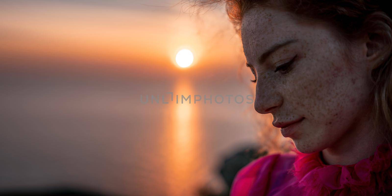 Close up portrait of curly redhead young caucasian woman with freckles looking at camera and smiling. Cute woman portrait in a pink long dress posing on a volcanic rock high above the sea at sunset by panophotograph