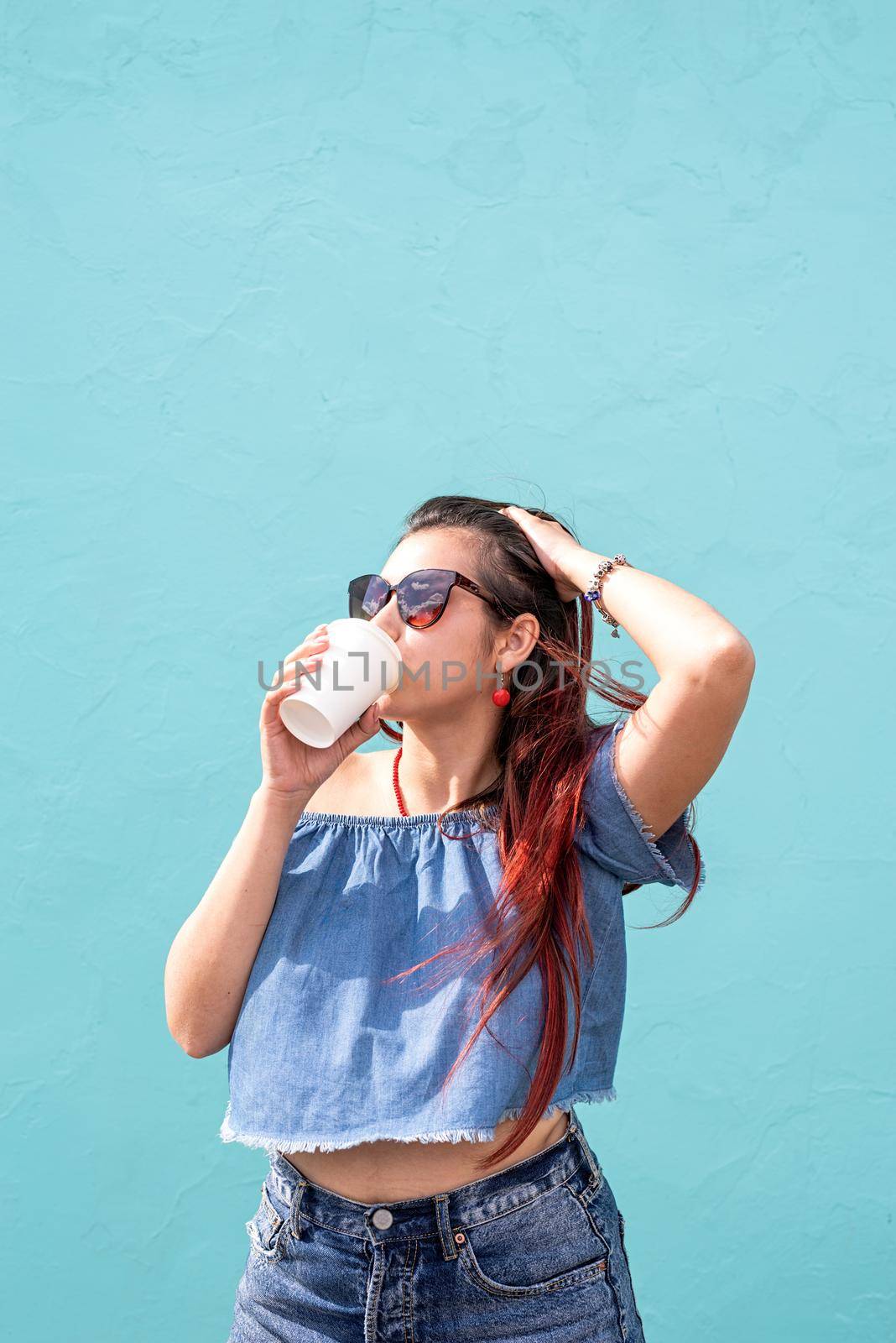 Attractive young woman in summer clothes and sunglasses holding cup of coffee in her hands, dancing on blue wall background at street