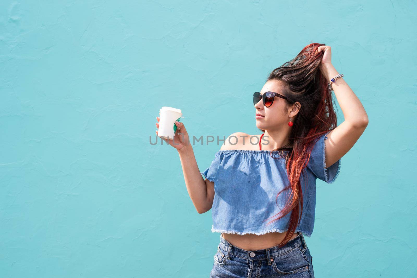 Attractive young woman in summer clothes and sunglasses holding cup of coffee in her hands, dancing on blue wall background at street