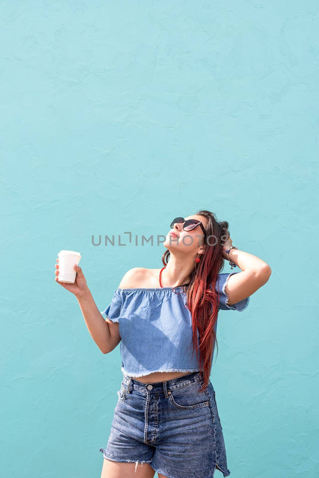 Attractive young woman in summer clothes and sunglasses holding cup of coffee in her hands, dancing on blue wall background at street