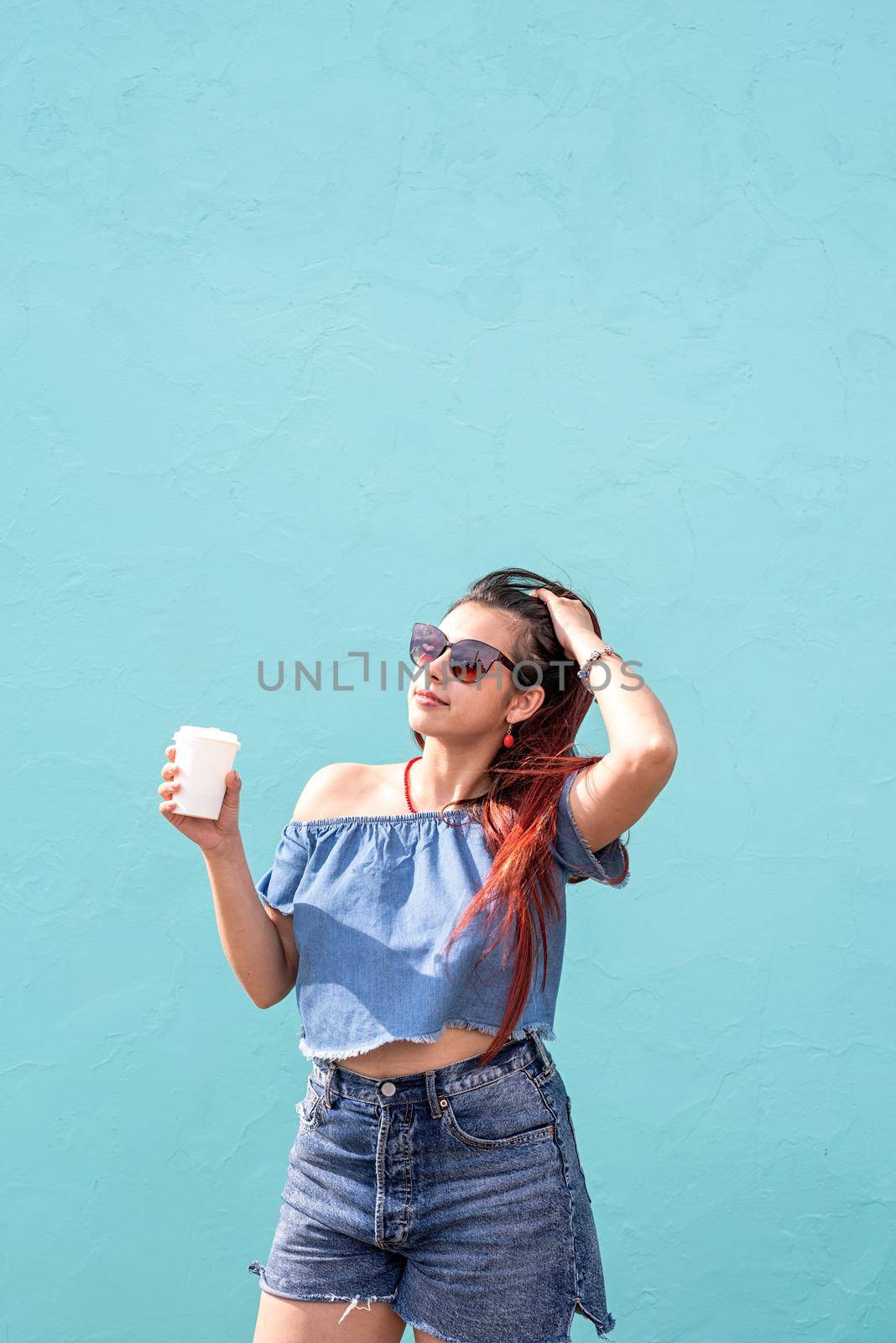 cheerful trendy woman with red hair drinking coffee dancing on blue wall background at street by Desperada