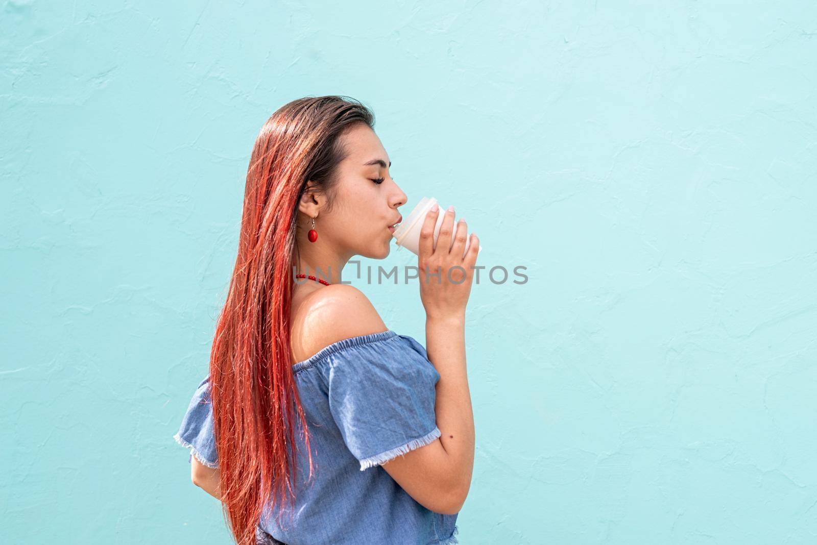 cheerful trendy woman with red hair drinking coffee dancing on blue wall background at street by Desperada