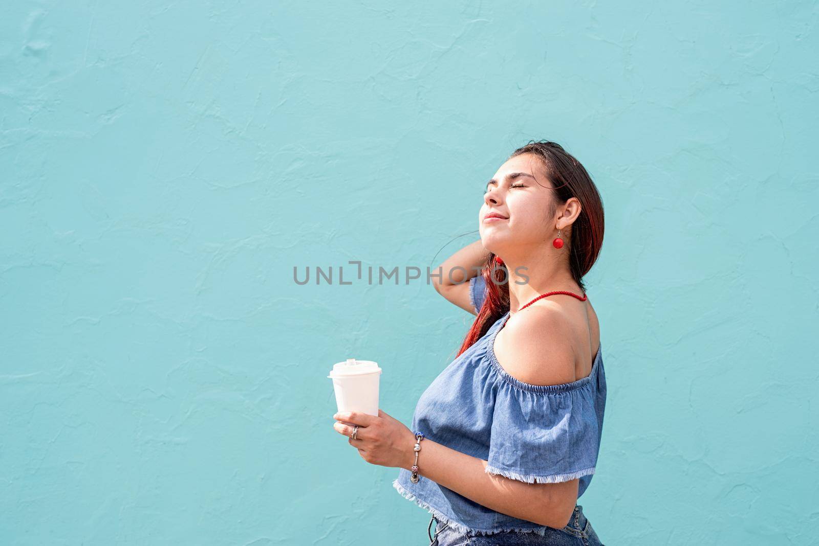 Attractive young woman in summer clothes and sunglasses holding cup of coffee in her hands, dancing on blue wall background at street