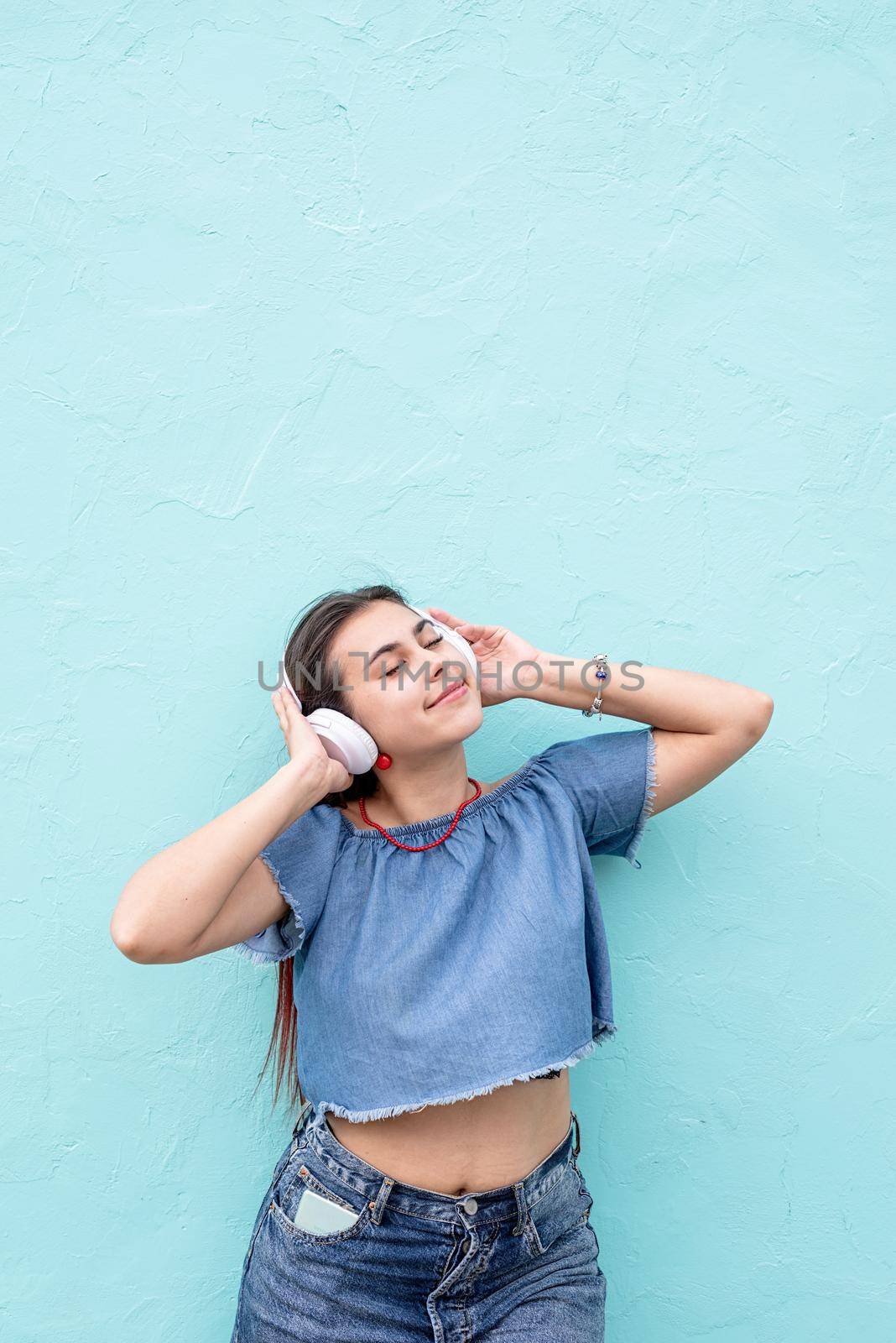 Attractive young woman in summer clothes and sunglasses listening to the music on blue wall background at street