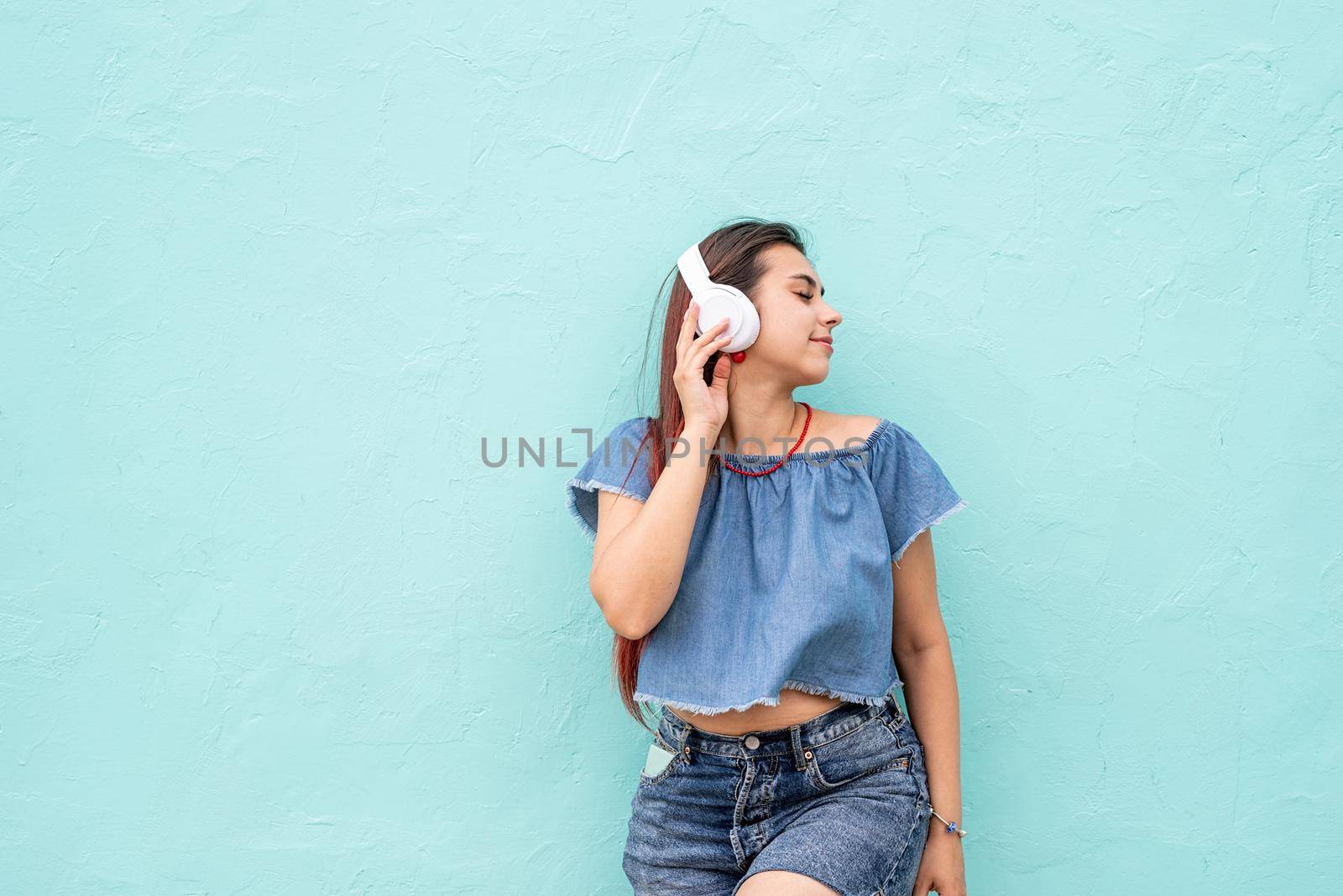 Attractive young woman in summer clothes and sunglasses listening to the music on blue wall background at street