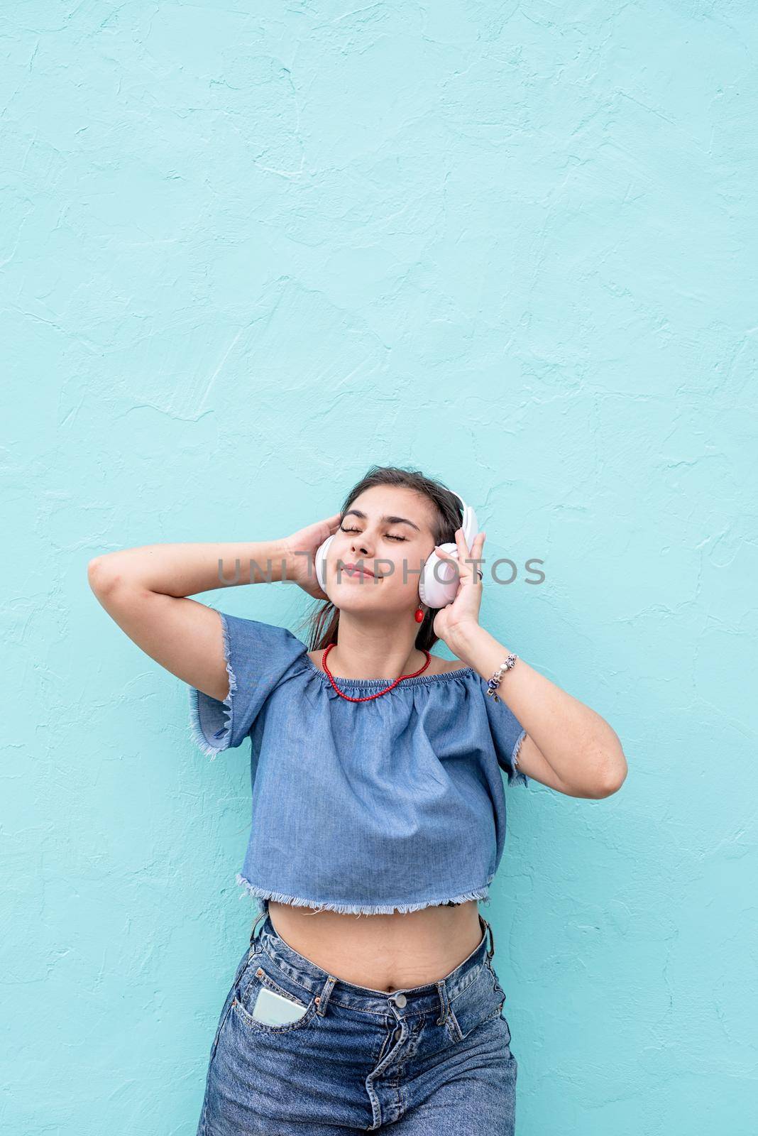 Attractive young woman in summer clothes and sunglasses listening to the music on blue wall background at street