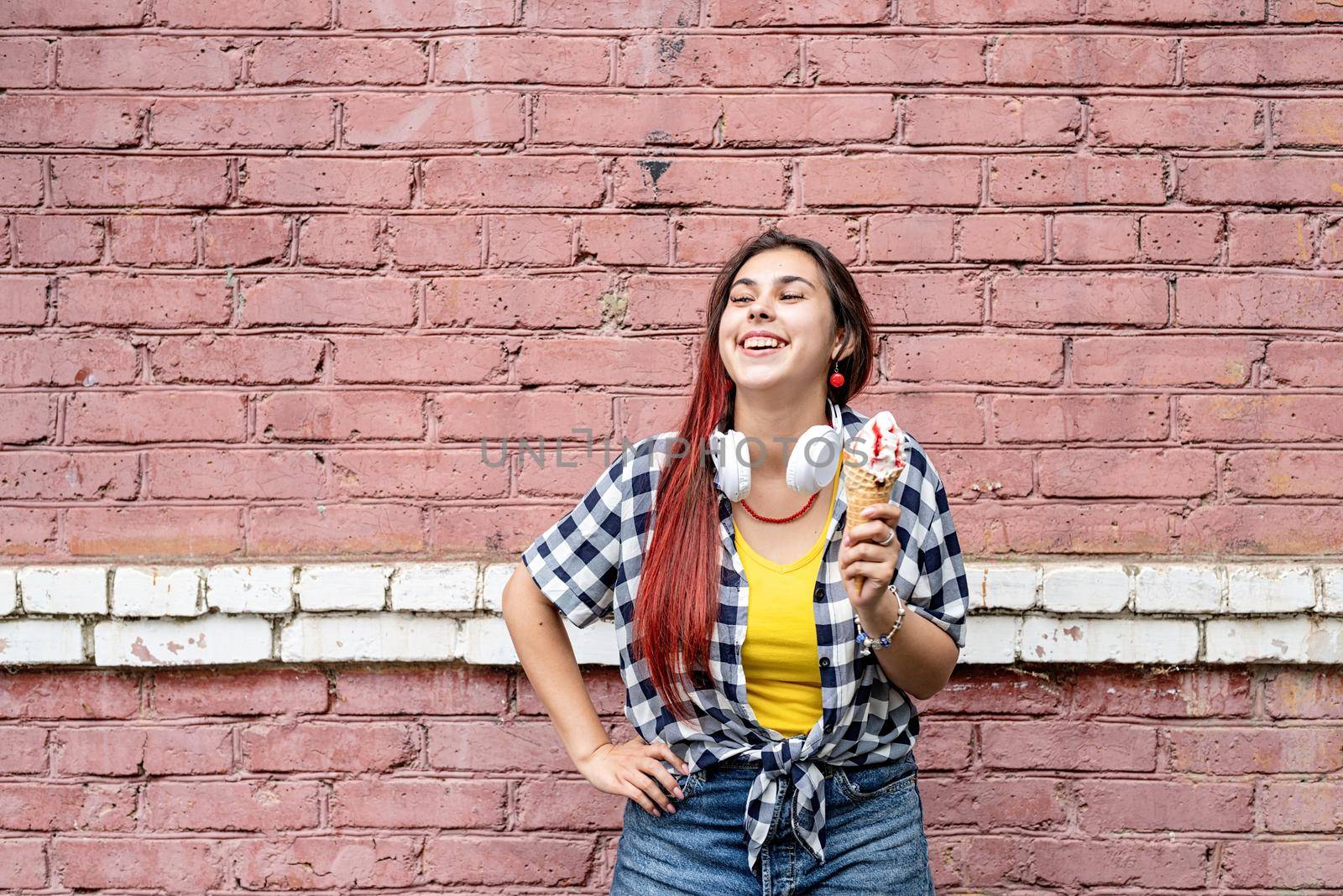 cheerful trendy woman with red hair eating ice cream on pink brick wall background at street by Desperada