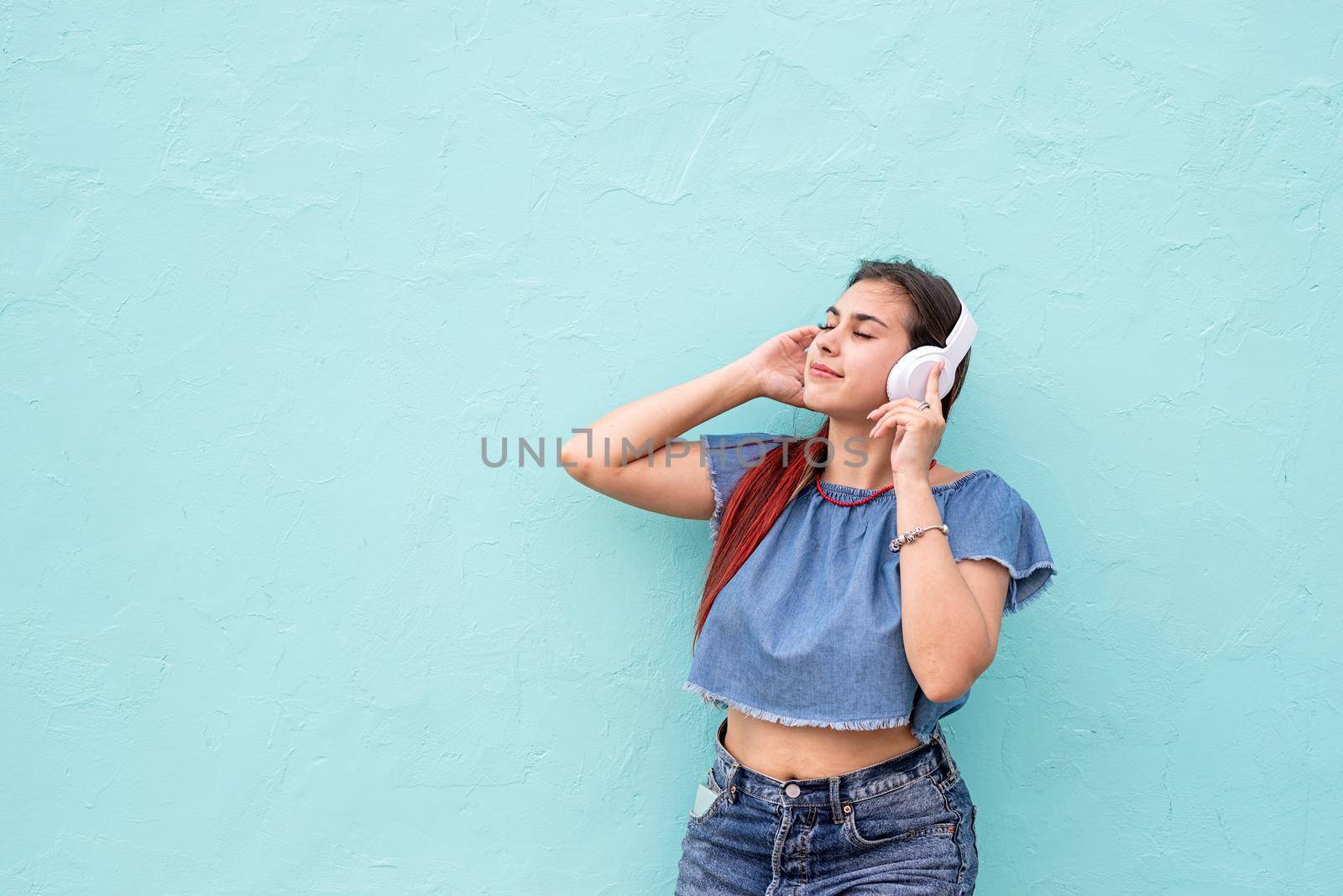 Attractive young woman in summer clothes and sunglasses listening to the music on blue wall background at street