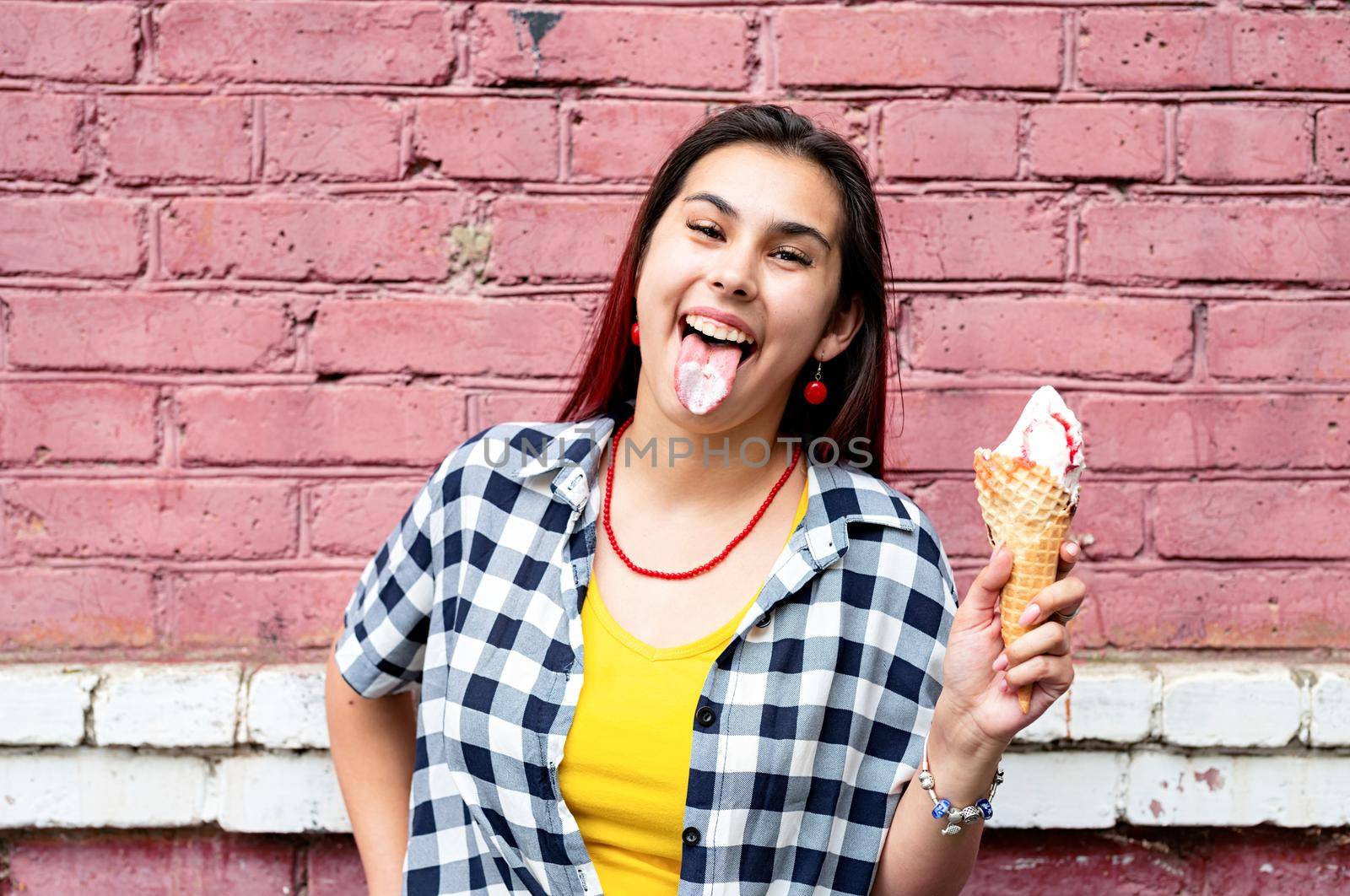 Attractive young woman in summer clothes and sunglasseseating ice cream on pink brick wall background at street