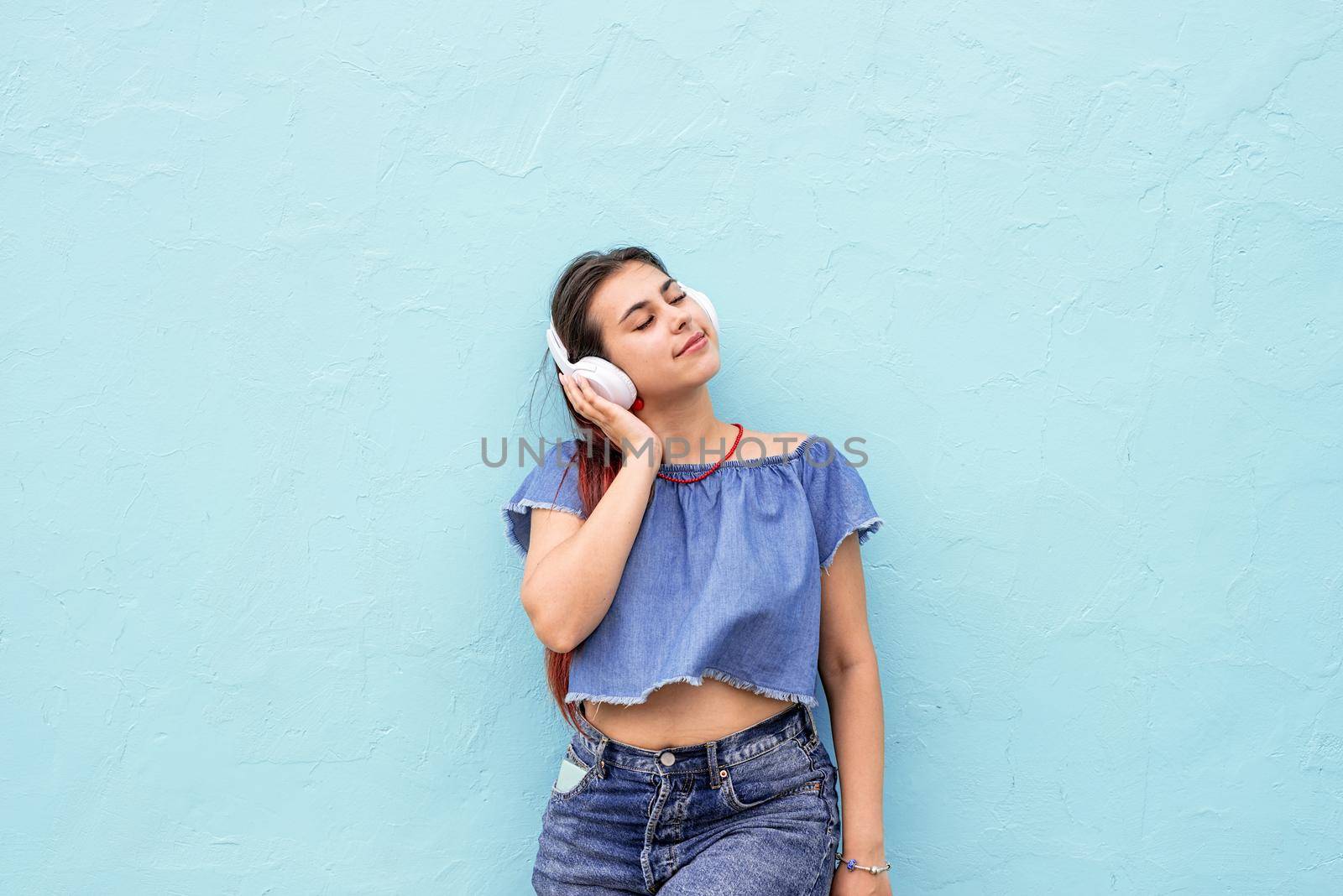 Attractive young woman in summer clothes and sunglasses listening to the music on blue wall background at street