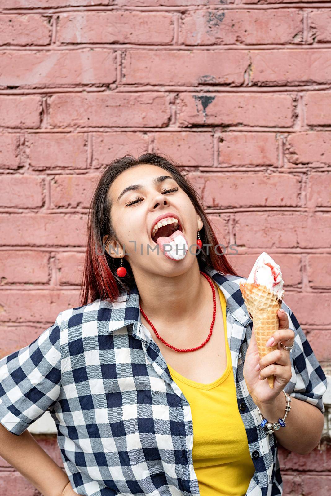 cheerful trendy woman with red hair eating ice cream on pink brick wall background at street by Desperada