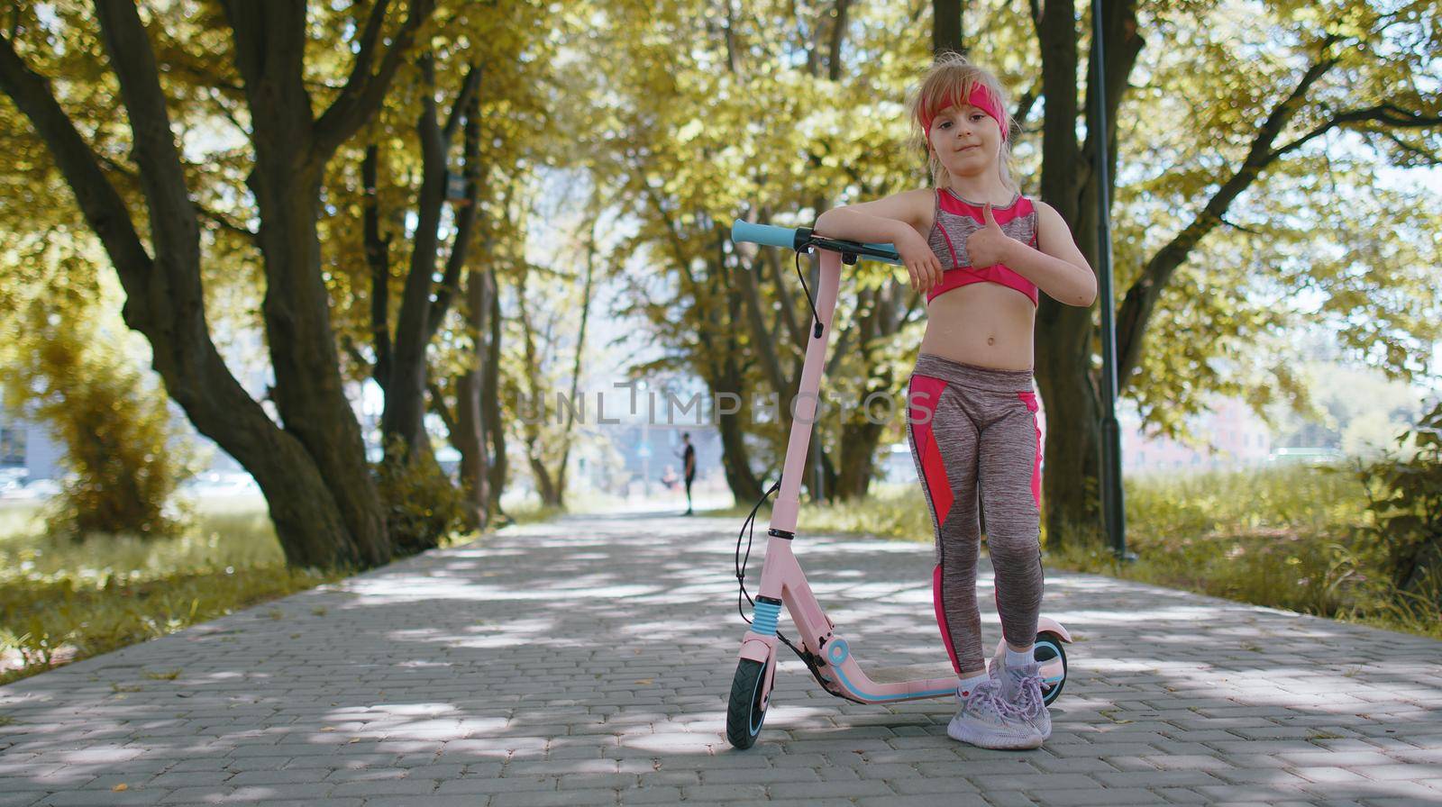 Young athletic fit blonde girl starts riding travel on electric scooter on road in park on sunny day. Child going outdoors, having fun. Kid leisure activity, sport. Modern technology for children