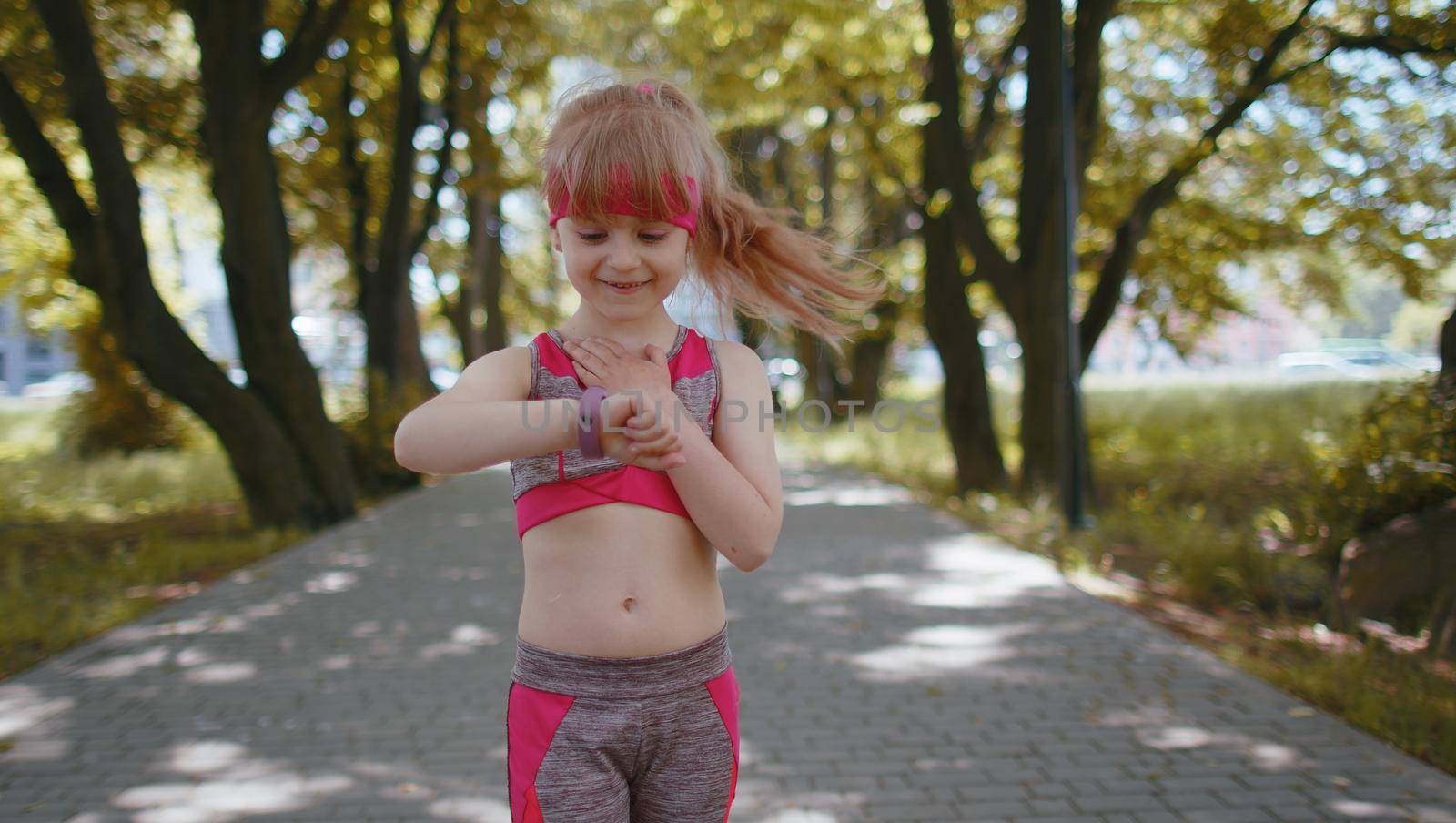 Athletic toddler runner girl look at smart watch clock device fitness bracelet checking time, heart rate, pulse, SPO2 on smartwatch on jogging. Workout cardio training outside in city park at morning