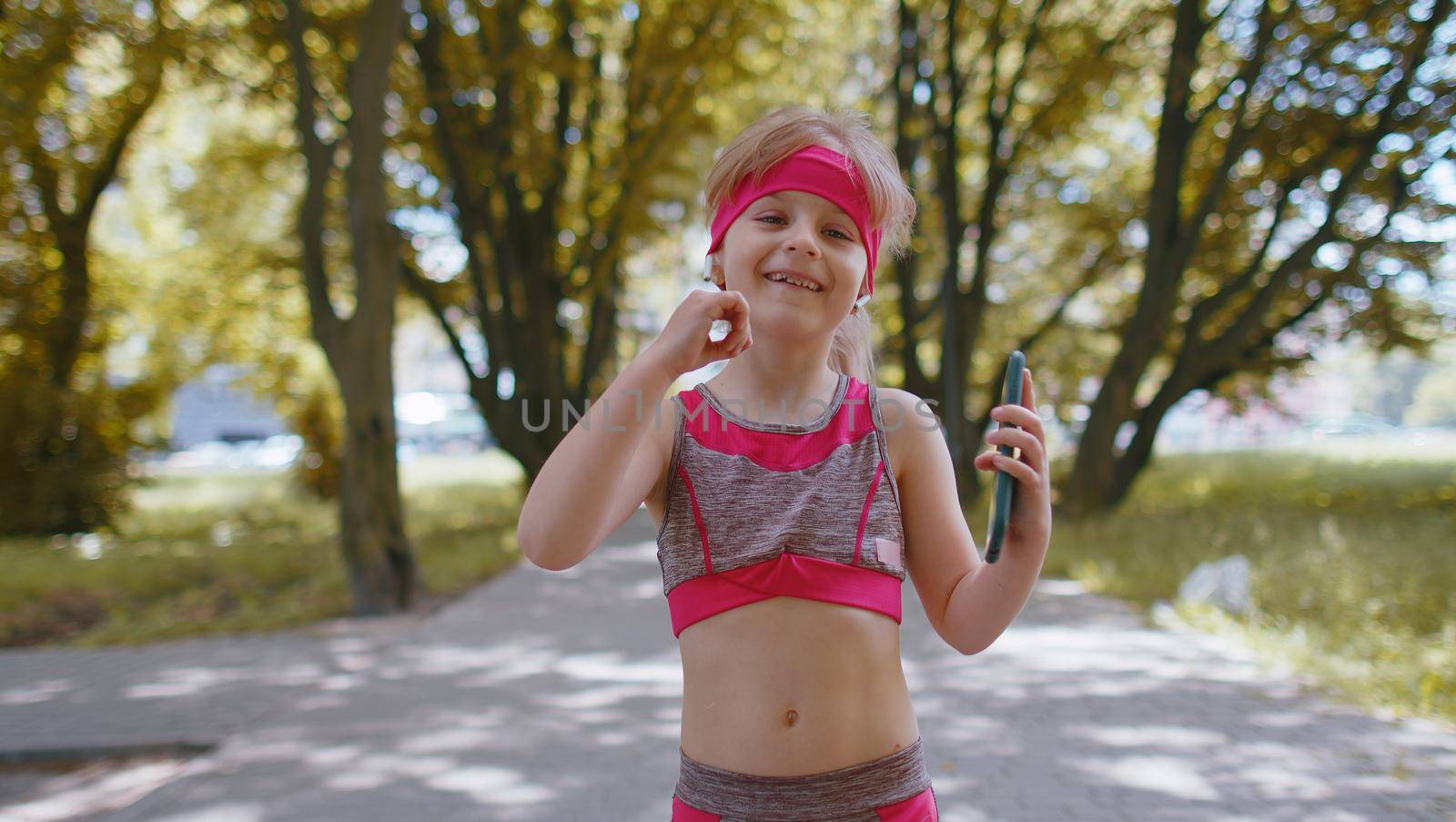 Athletic fitness sport runner child girl walking and using smartphone listening music with wireless earphones. Workout cardio in morning park. Children toddler kid jogger enjoying healthy lifestyle