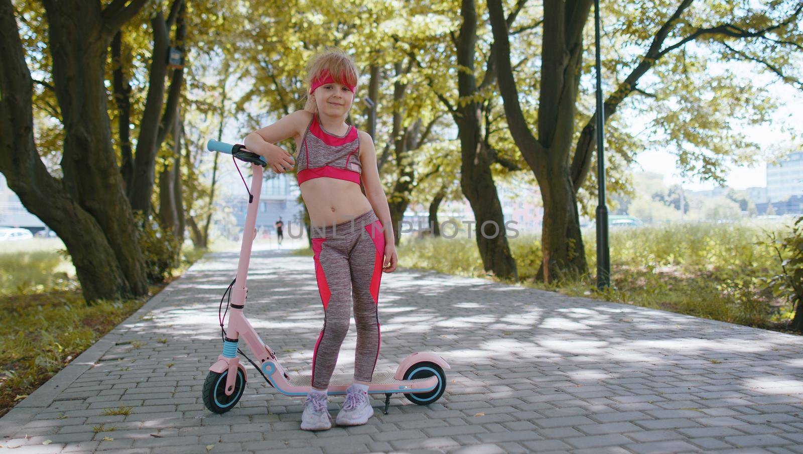 Athletic fit young girl rides on electric scooter on road in park on sunny day, outdoors. Child going standing near bike looking at camera. Kid leisure activity, sport. Modern technology for children