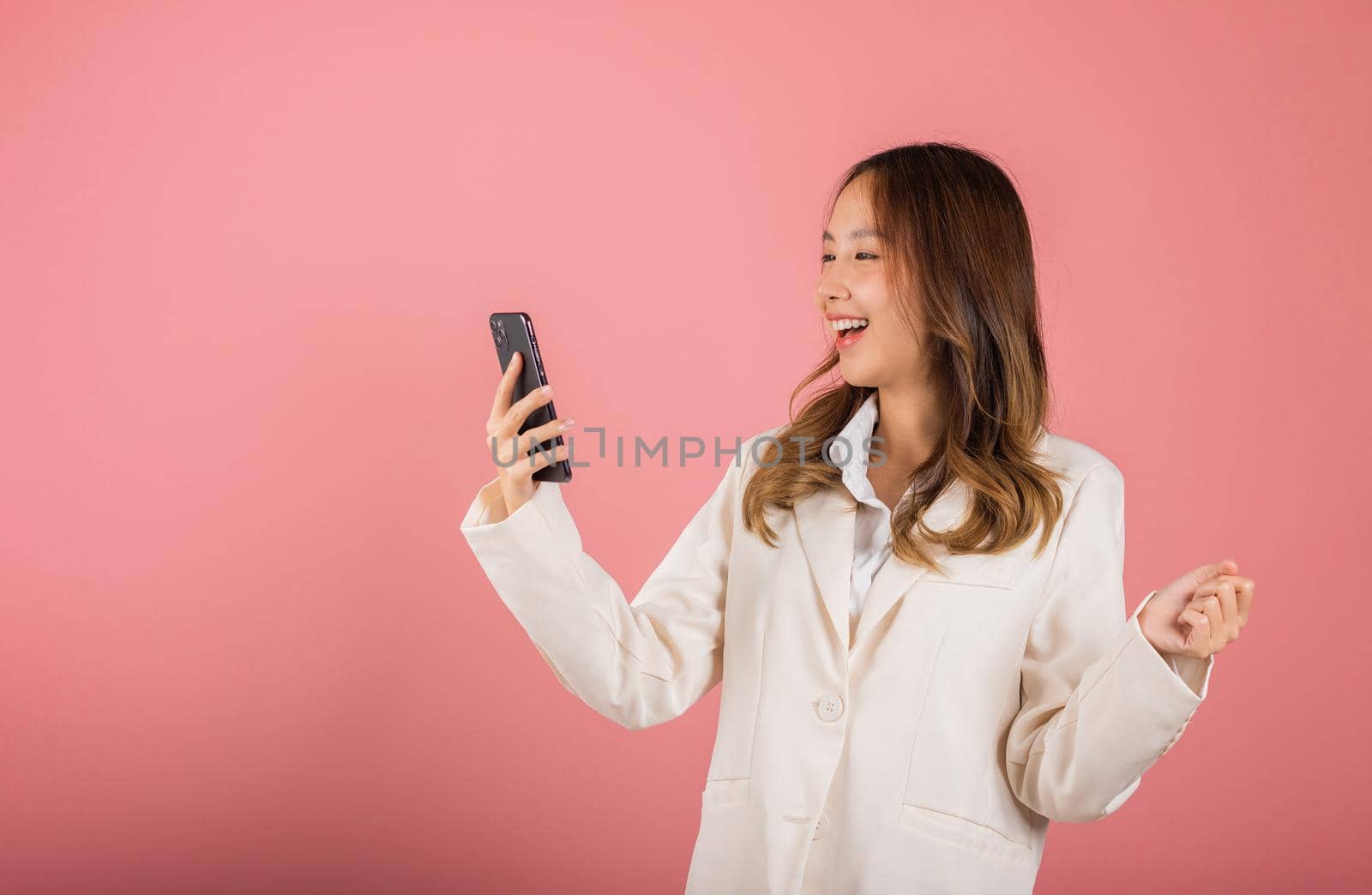 Happy Asian portrait beautiful cute young woman teen smiling excited face using mobile phone say yes! Studio shot isolated on pink background, Thai female surprised make winner gesture on smartphone