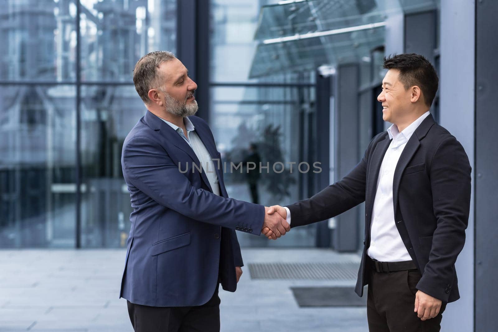 Two businessmen, bosses greet each other by shaking hands, a diverse group of men in business clothes outside an office building