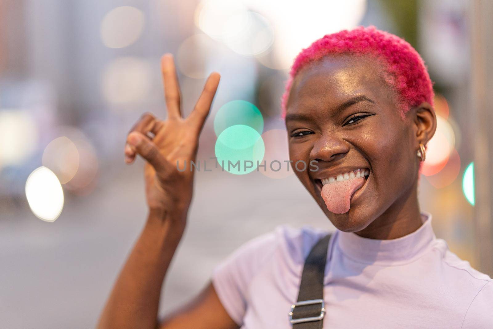 African woman with short pink hair grimacing at camera gesturing victory with two fingers outdoors
