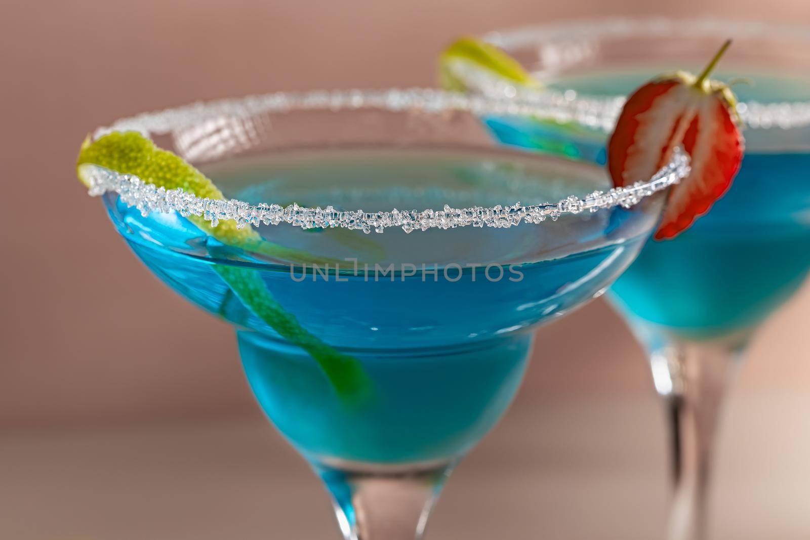 Two glasses with blue margarita cocktail garnished with lime zest and strawberries, selective focus.